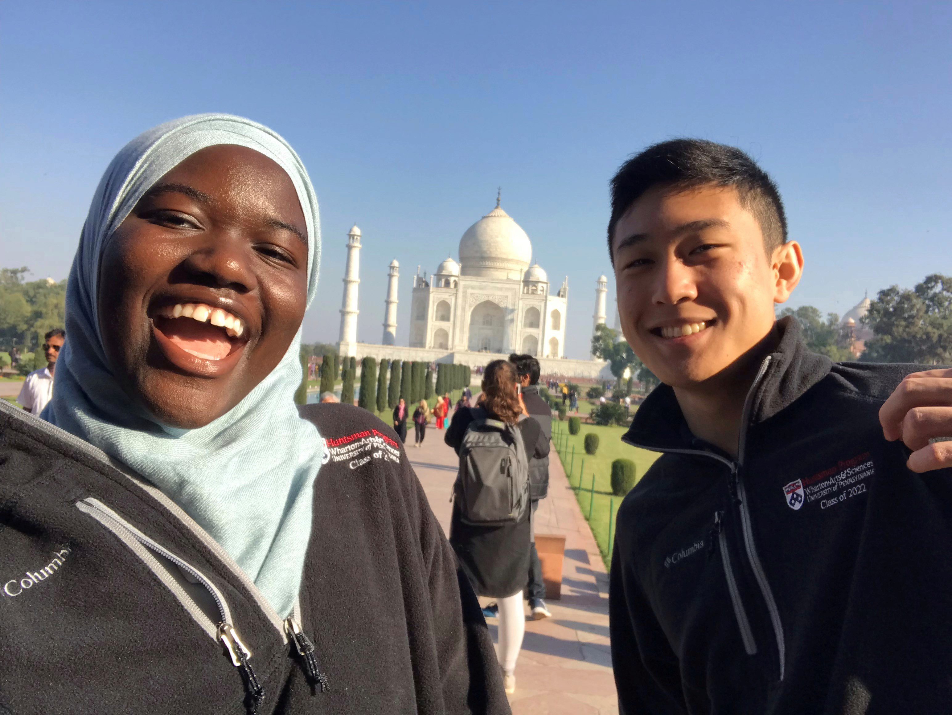 Two people are smiling in front of the Taj Mahal, a famous white marble mausoleum in Agra, India. They are wearing jackets with university logos.