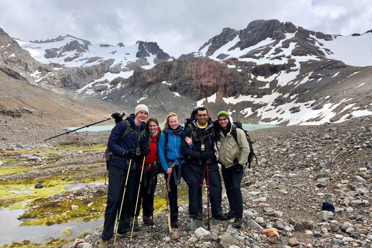 A group of hikers poses on a rocky mountain landscape with snow patches and a glacier-fed lake in the background. They are dressed in cold-weather gear.