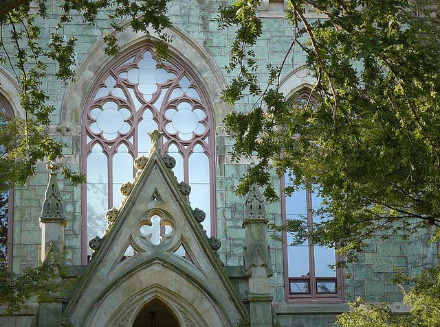 Gothic-style architecture with arched windows, intricate tracery designs, and stone details, partially obscured by tree branches.