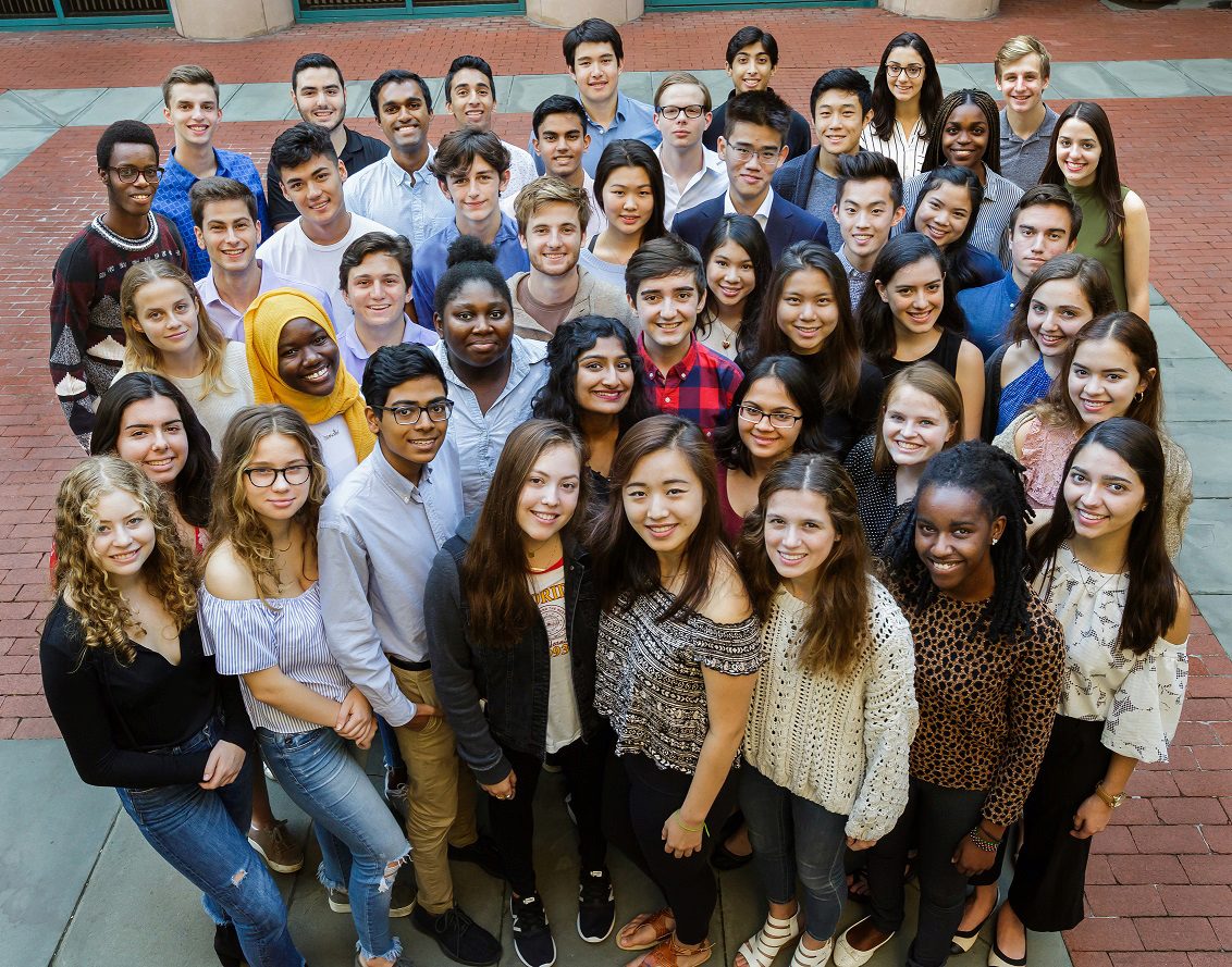 A diverse group of young adults standing closely together outside on a brick-paved area, smiling and looking up at the camera.