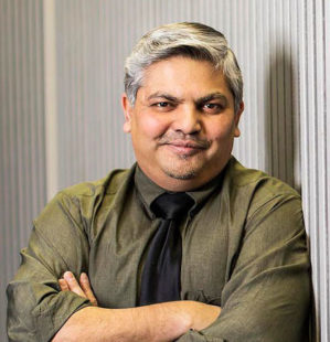 Headshot of a person with gray hair, wearing a dark green shirt and black tie, standing with folded arms against a striped backdrop.