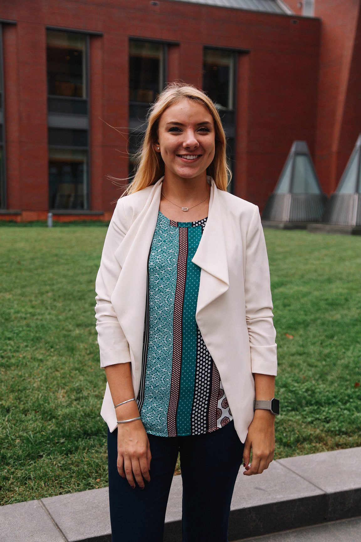 A person smiling outdoors, wearing a white blazer over a patterned blouse, standing in front of a building with large windows.