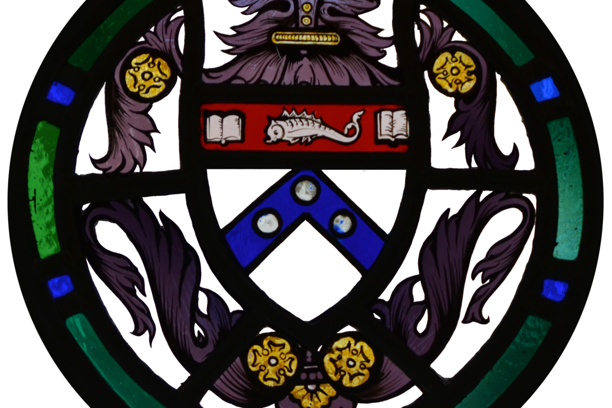 Stained glass circular design featuring a coat of arms with a chevron, three roundels, and two open books. Flanked by ornate patterns and a sea creature motif.