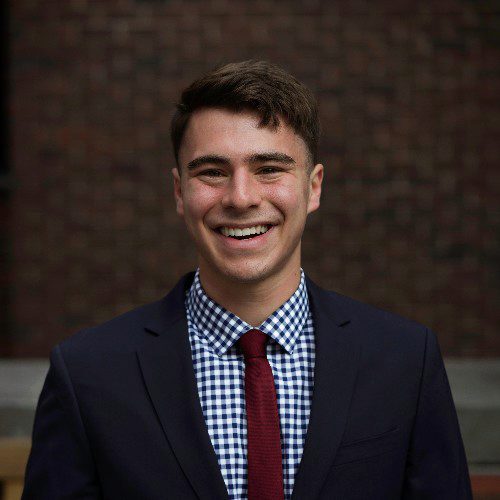 Headshot of a person wearing a dark suit, checkered shirt, and red tie, smiling with a blurred brick wall background.