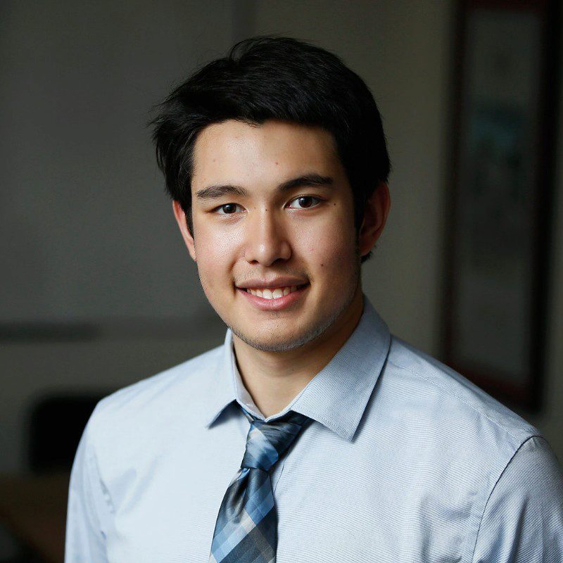 A professional headshot of a person wearing a light blue shirt and a blue patterned tie, smiling at the camera.