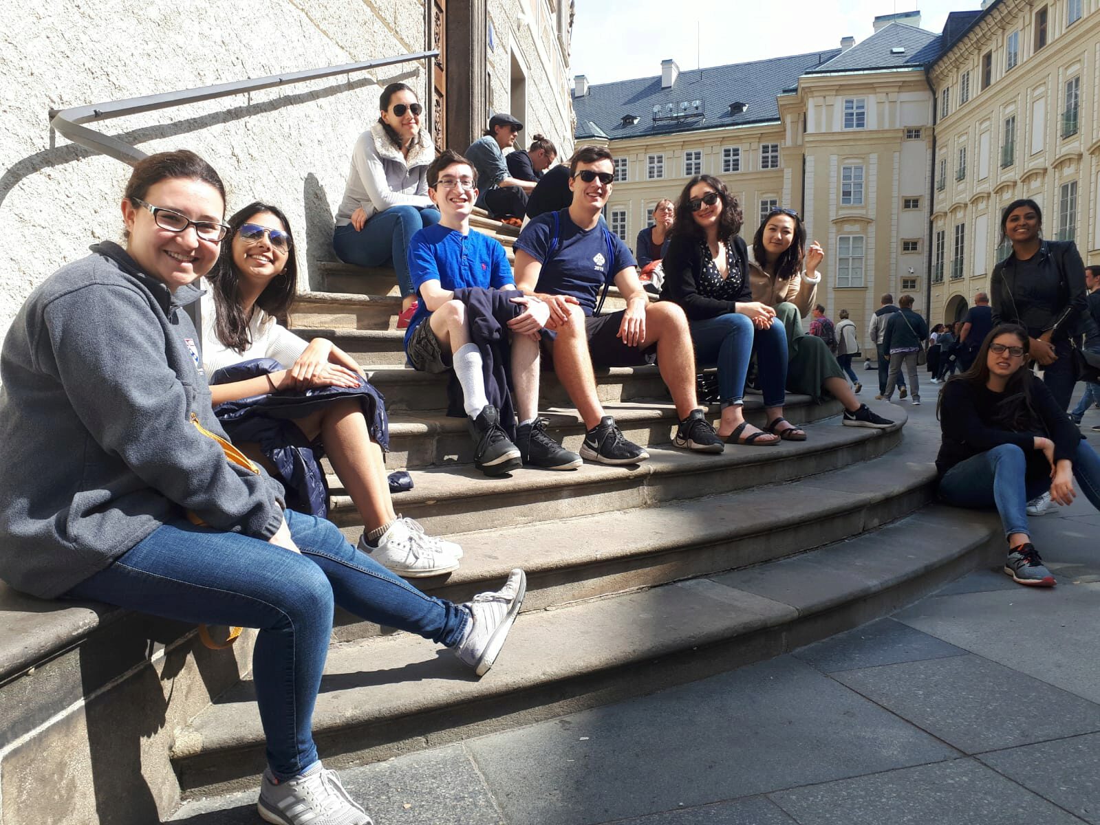 A group of people sitting and smiling on outdoor stone steps in a sunny courtyard with historic buildings in the background.