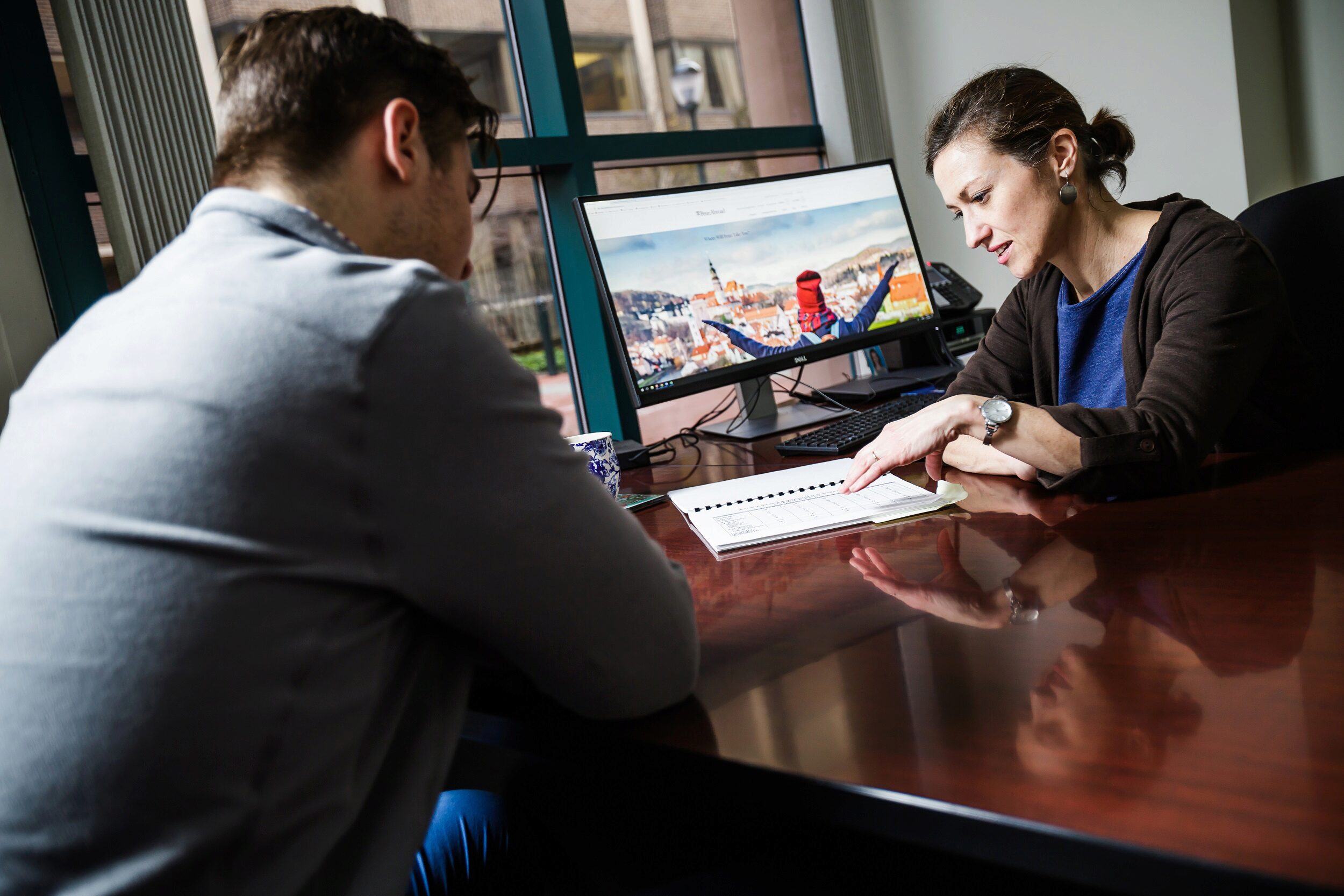 Two people are sitting at a desk in an office, with one person pointing at a document. A computer screen displays a scenic landscape.