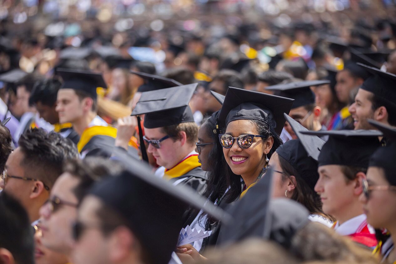 A large group of graduates wearing caps and gowns seated at a graduation ceremony, with one smiling individual prominently in the foreground.