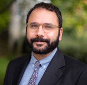 Headshot of a person with glasses, a beard, and mustache, wearing a suit jacket, dress shirt, and patterned tie, set against a blurred outdoor background.