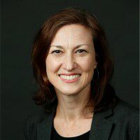 A professional headshot of a smiling person with shoulder-length brown hair, wearing a dark blazer and a black top.