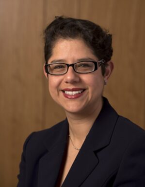 Headshot of a person with short black hair, wearing glasses, a dark blazer, and a necklace, smiling against a wooden background.