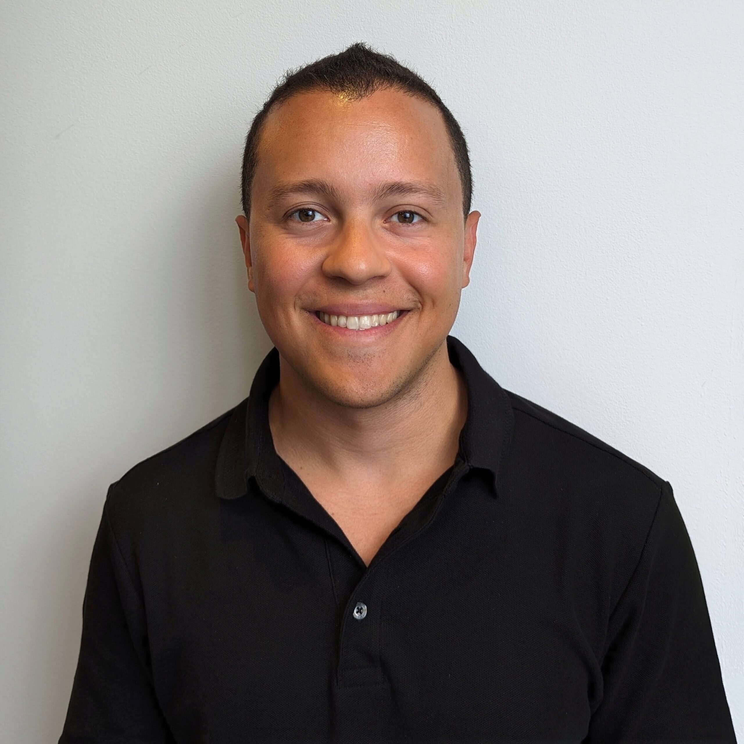 A headshot of a person wearing a black collared shirt, smiling in front of a plain white background.