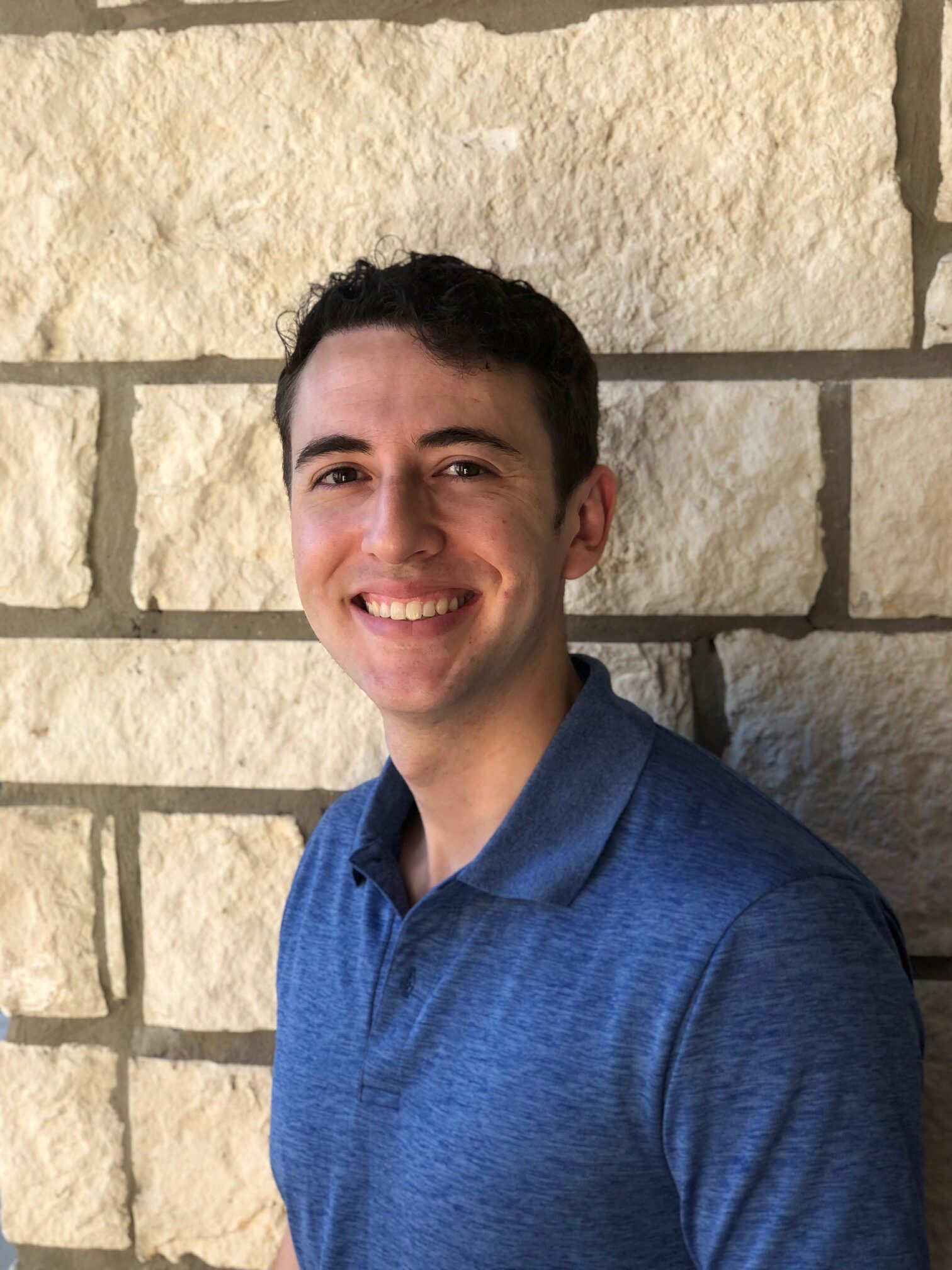 Headshot of a smiling person wearing a blue polo shirt in front of a stone wall.