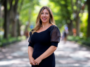 A person with long hair wearing a black dress stands on a tree-lined path on a sunny day, smiling at the camera.