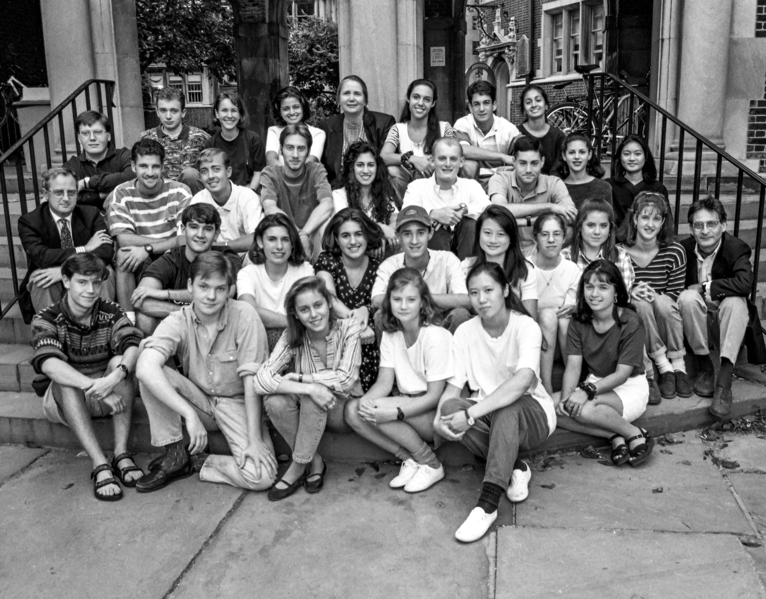 A black and white group photo of people sitting on outdoor steps, likely a class or team. The group is diverse and poses casually, some smiling at the camera.