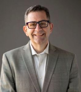 A person wearing glasses and a light gray suit jacket over a white shirt, smiling in a headshot against a neutral background.