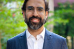 A headshot of a person with glasses and a beard, wearing a dark suit and white shirt, smiling outdoors with greenery in the background.