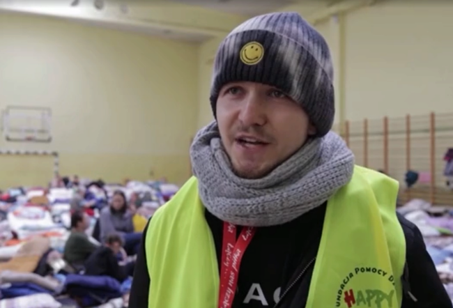 A person wearing a beanie with a smiley face and a scarf, dressed in a yellow safety vest, is in a crowded indoor space. The room is filled with people and bedding items, indicating a temporary shelter or assistance setup.