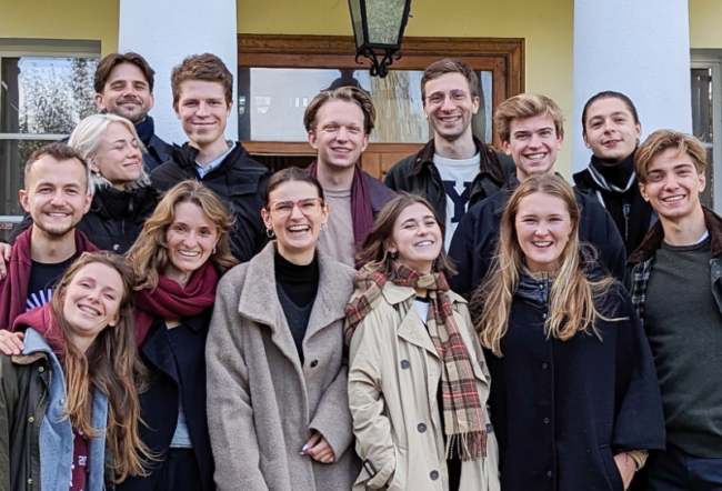 A group of thirteen people smiling in an outdoor setting, standing closely together in front of a building with white columns and a wooden door.