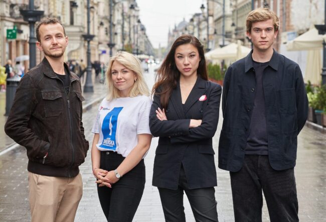 Four people standing on an urban street, posing confidently. The background shows a cityscape with buildings and people in the distance.