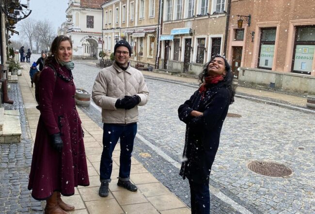 Three people standing on a cobblestone street in a European-style city, surrounded by lightly snowing weather and historic buildings.