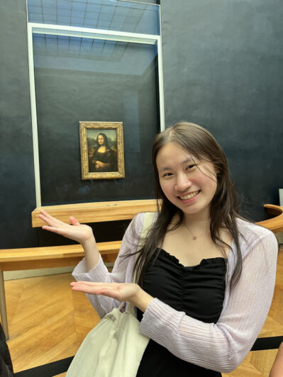 A person smiling and gesturing towards the Mona Lisa painting in a museum setting.